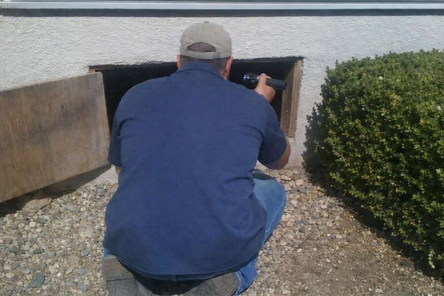 A man kneeling at the open crawl-space access of a house during an inspection